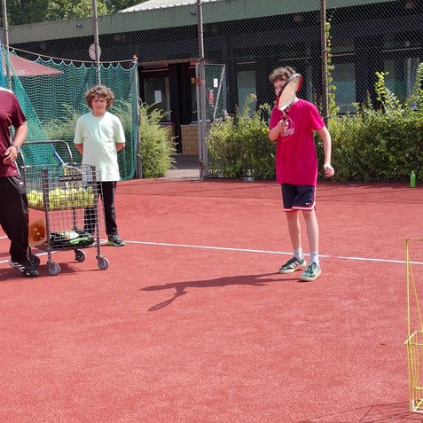 Zwei Kinder stehen auf einem Tennisplatz, w&auml;hrend ein Junge mit Schl&auml;ger zur Schlagbewegung ansetzt.