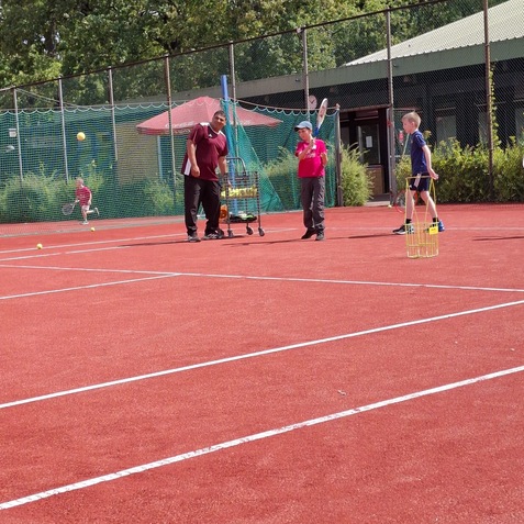 Trainingssituation auf einem roten Tennisplatz mit einem Trainer und drei Kindern, die Tennis spielen und &uuml;ben.