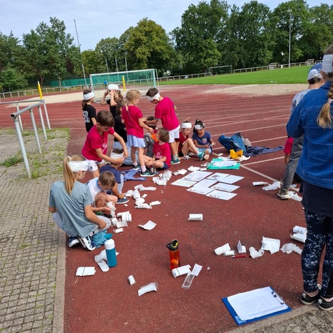 Gruppe von Kindern in Rot, die auf einem Sportplatz mit Materialien hantieren, umgeben von Papier und Notizen.