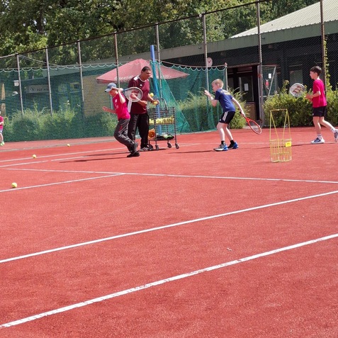 Trainingsszene auf einem roten Tennisplatz mit Kindern, die Tennis spielen, und einem Trainer mit B&auml;llen.