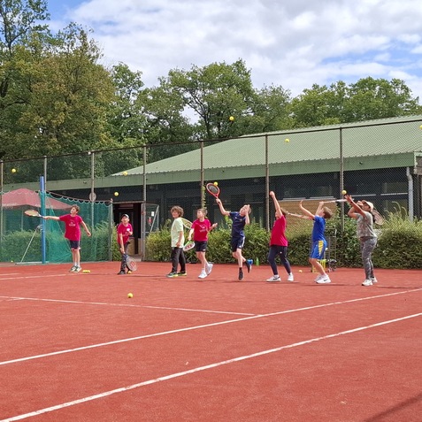 Gruppe von Kindern beim Tennistraining auf einem roten Platz, Schl&auml;ger in der Hand, B&auml;lle fliegen durch die Luft.