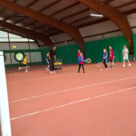 Gruppe von Kindern in einem Tennisunterricht, die auf einem Hallenplatz mit B&auml;llen und Schl&auml;gern trainieren.
