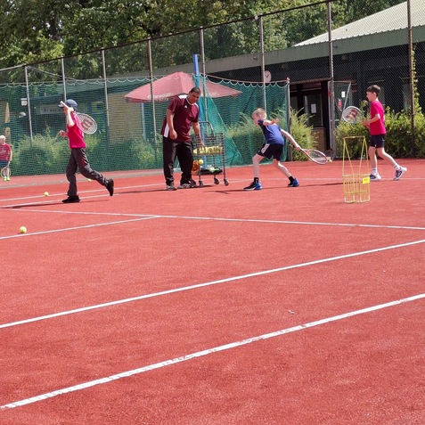 Vier Kinder &uuml;ben Tennis auf einem roten Platz, w&auml;hrend ein Trainer ihnen Anweisungen gibt und B&auml;lle bereitstellt.