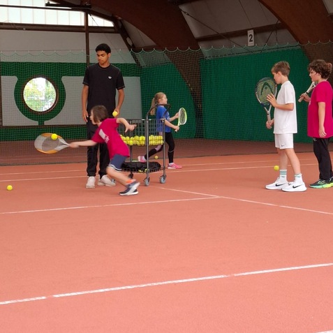 Gruppe von Kindern beim Tennistraining in einer Halle, ein Trainer beobachtet sie, B&auml;lle liegen in einem Wagen.