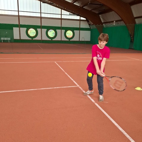 Junge mit Tennisschl&auml;ger schl&auml;gt einen Ball auf einem Tennisplatz mit rotem Belag und gr&uuml;nem Hintergrund.