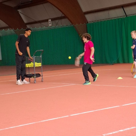 Zwei Kinder üben Tennis in einer Halle, ein Trainer steht neben einem Wagen mit Tennisbällen.