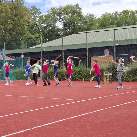 Gruppe von Kindern mit Tennisschl&auml;gern beim Training auf einem roten Tennisplatz unter einem bew&ouml;lkten Himmel.