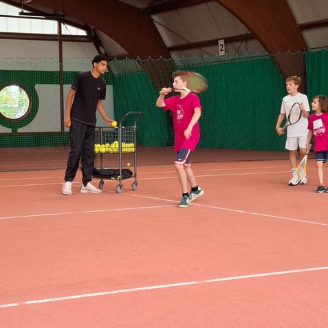 Kinder trainieren Tennis in einer Sporthalle, w&auml;hrend ein Trainer B&auml;lle aus einem Wagen reicht.