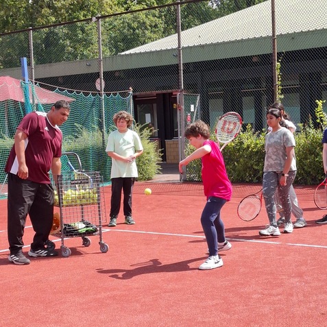 Trainer zeigt Kindern das Tennisspielen auf einem roten Platz, w&auml;hrend ein Ballwagen bereitsteht.
