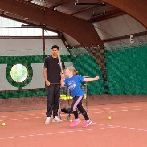 Ein M&auml;dchen in blauer T-Shirt springt auf einem Tennisplatz, w&auml;hrend ein Trainer beobachten und B&auml;lle daneben h&auml;lt.