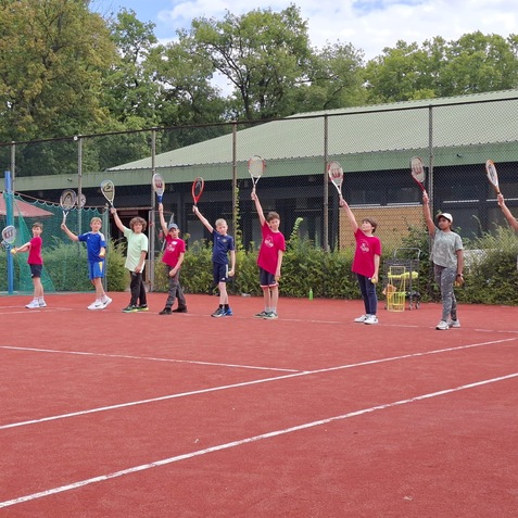 Gruppe von Kindern und einem Trainer auf einem Tennisplatz hebt ihre Schl&auml;ger zum Training in die H&ouml;he.