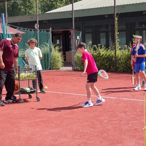 Vier Kinder trainieren auf einem Tennisplatz, dabei steht ein Trainer mit einem Wagen voller Tennisb&auml;lle.