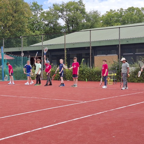 Gruppe von Kindern in Sportbekleidung mit Tennisschl&auml;gern auf einem roten Tennisplatz, begleitet von einem Trainer.