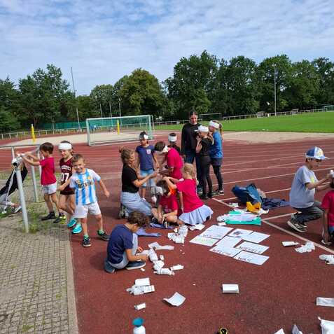 Gruppe von Kindern und Erwachsenen auf einem Sportplatz, einige sitzen, andere stehen und basteln gemeinsam.