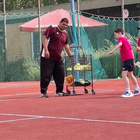 Trainer mit Wagen voll Tennisb&auml;llen auf rotem Platz, w&auml;hrend zwei Kinder &uuml;ben. Schatten eines Schirms im Hintergrund.