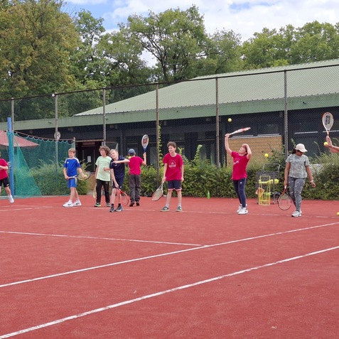 Gruppe von Kindern auf einem Tennisplatz, die mit Schl&auml;gern B&auml;lle schlagen und Tennis&uuml;bungen machen.