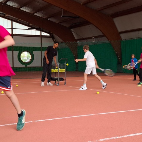 Eine Tennis&uuml;bung mit Kindern in einem Hallenplatz. Ein Trainer steht neben einem Wagen mit Tennisb&auml;llen.