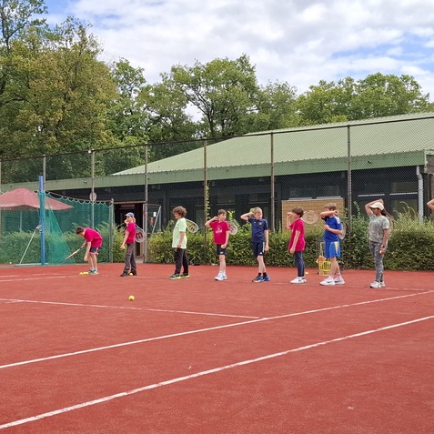 Sechs Kinder stehen auf einem roten Tennisplatz, einige b&uuml;cken sich, andere schauen aufmerksam, B&auml;ume im Hintergrund.