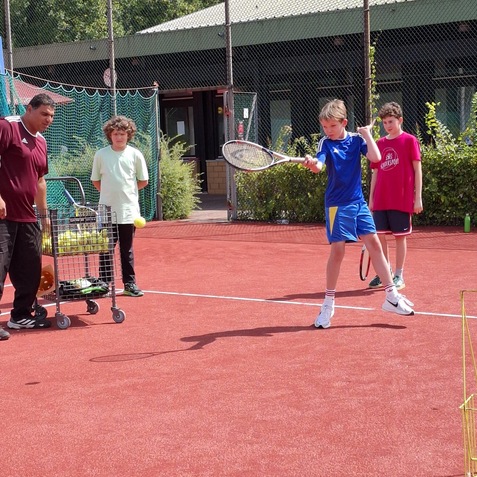 Junge mit Tennisschl&auml;ger schl&auml;gt einen Ball auf einem roten Tennisplatz, Trainer und zwei Kinder beobachten.