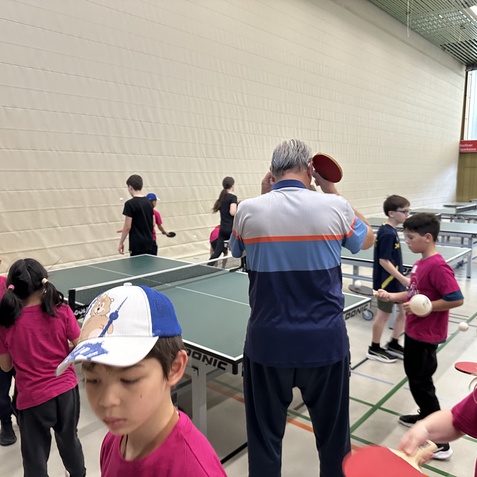 Gruppe von Kindern in Trainingskleidung mit Tischtennisschlägern in einer Halle mit mehreren Tischtennisplatten.