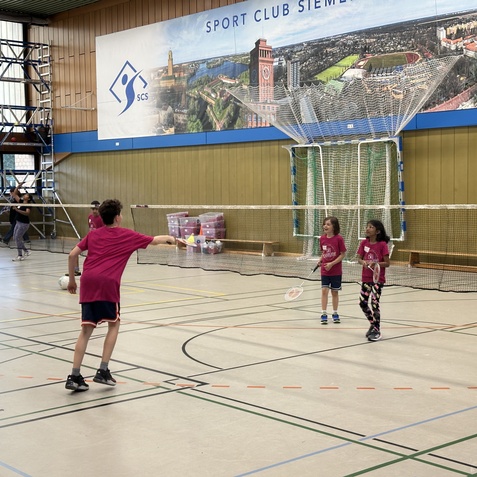 Drei Kinder in pinken T-Shirts spielen Badminton in einer Sporthalle, im Hintergrund ist eine Tribüne sichtbar.