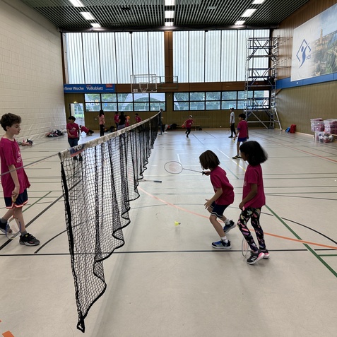 Kinder in pinken T-Shirts spielen Badminton in einer Sporthalle mit Netzen und verschiedenen Sportgeräten im Hintergrund.