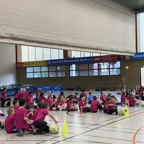 Gruppe von Kindern in pinken T-Shirts sitzt auf dem Boden einer Sporthalle, umgeben von Bällen und Markierungskegeln.