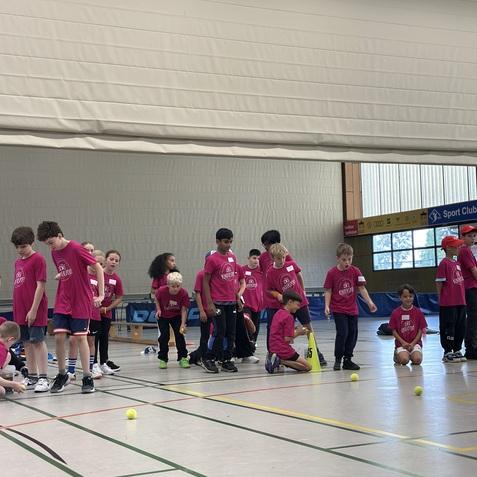 Gruppenspiel mit Kindern in pinken T-Shirts, die auf einem Hallenboden mit Tennisbällen interagieren.