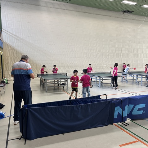 Kinder in pinken Shirts spielen Tischtennis in einer Sporthalle, ein Trainer beobachtet sie am Rand.