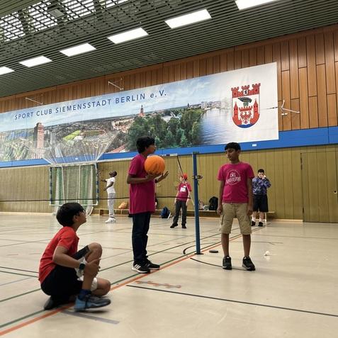 Zwei Kinder in pinken T-Shirts spielen mit einem Basketball in einer Sporthalle, in der anderen Kinder aktiv sind.