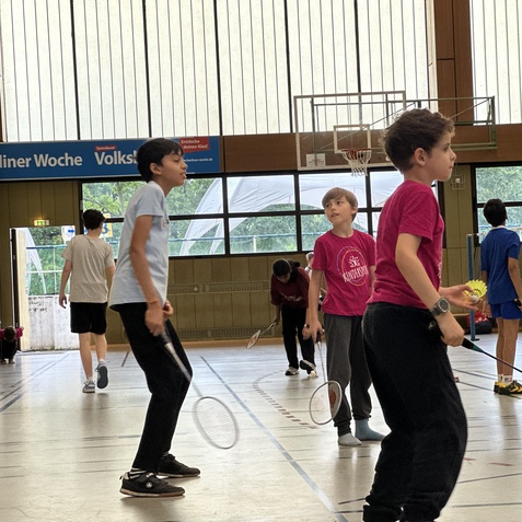 Gruppe von Kindern in Sportkleidung beim Badmintonspielen in einer Halle mit Fenster und Sportger&auml;ten.