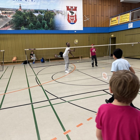 Kind und Jugendliche spielen Badminton in einer Sporthalle mit Netz und Linien auf dem Boden.