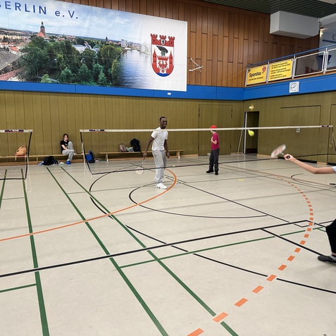Zwei Jugendliche spielen Badminton in einer Sporthalle mit Netzen und einem Wandbild im Hintergrund.
