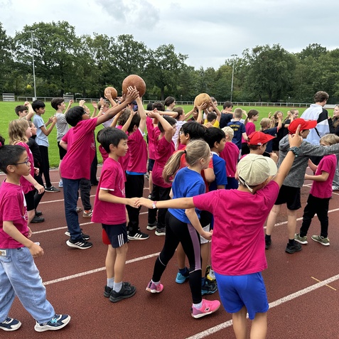 Gruppenspiel mit Kindern in pinken T-Shirts und B&auml;llen auf einer Laufbahn, umgeben von gr&uuml;ner Wiese und B&auml;umen.