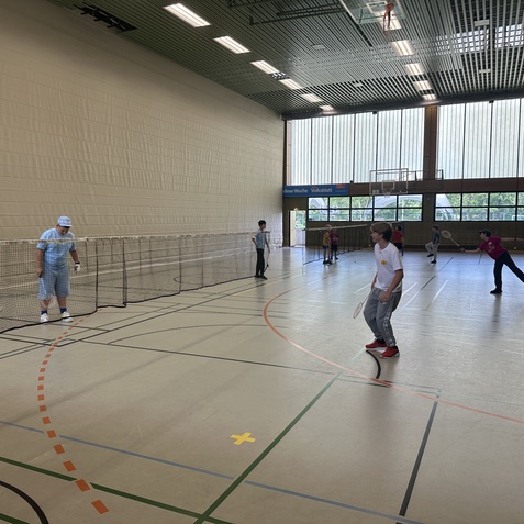 Jugendliche spielen Badminton in einer gro&szlig;en Sporthalle mit Netzen und hellem Tageslicht.