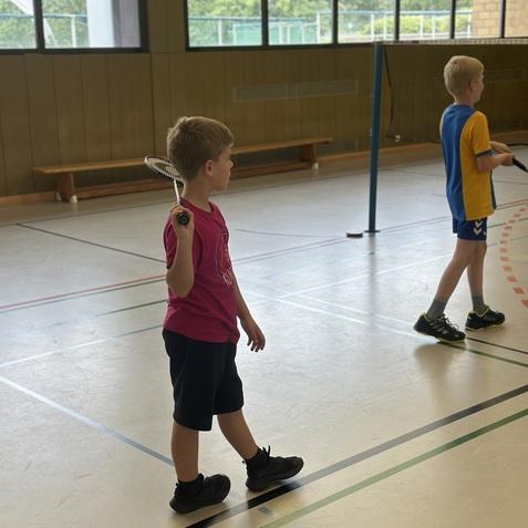 Ein Junge in pinkem T-Shirt h&auml;lt einen Badmintonschl&auml;ger, w&auml;hrend ein weiterer Junge auf einem Sportboden steht.
