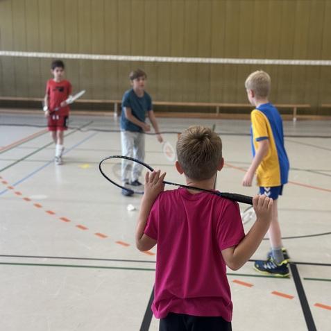 Vier Kinder spielen Badminton in einer Turnhalle, wobei ein Junge mit einem Schl&auml;ger im Vordergrund steht.