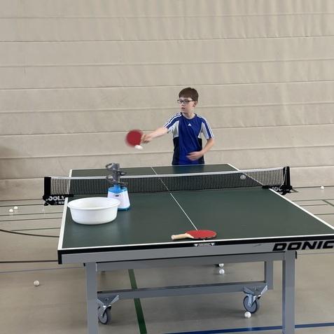 Junge mit Brille schl&auml;gt einen Tischtennisball mit einem Schl&auml;ger auf einem Tischtennistisch in einer Sporthalle.