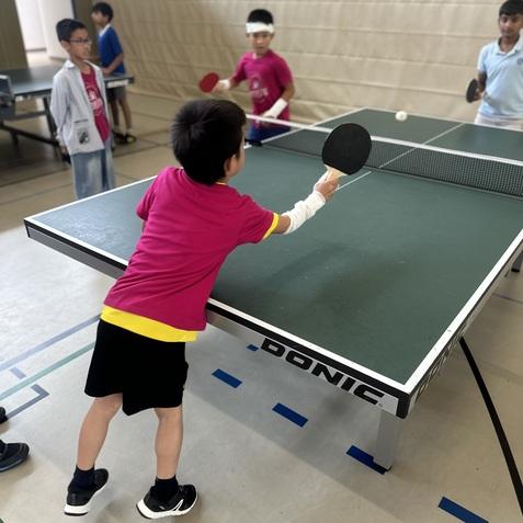 Kinder spielen Tischtennis in einer Halle; ein Junge schl&auml;gt den Ball mit einem Schl&auml;ger auf einer gr&uuml;nen Tischtennisplatte.
