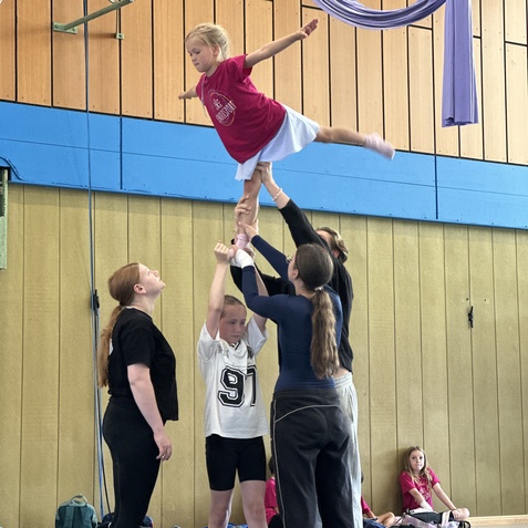 M&auml;dchen balanciert mit ausgestreckten Armen auf den H&auml;nden von vier anderen Kinder in einer Turnhalle.