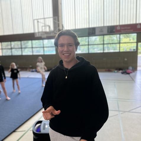 Junger Mann in schwarzer Jacke steht l&auml;chelnd in einer Sporthalle, im Hintergrund sind Personen auf einer Matte.