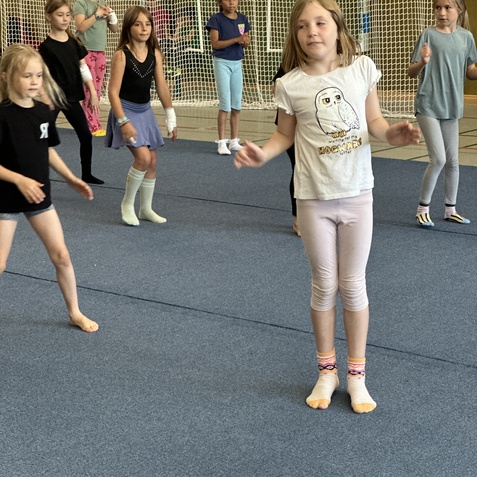 M&auml;dchen tanzen in einer Turnhalle auf blauen Matten, einige tragen bunte Socken und T-Shirts.