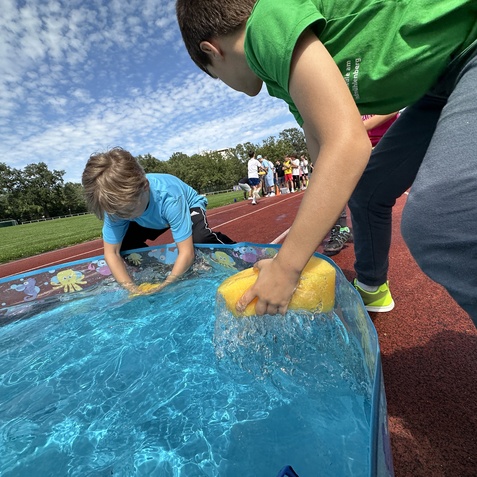 Zwei Kinder reinigen mit Schw&auml;mmen ein aufblasbares Schwimmbecken auf einem Sportplatz unter blauem Himmel.