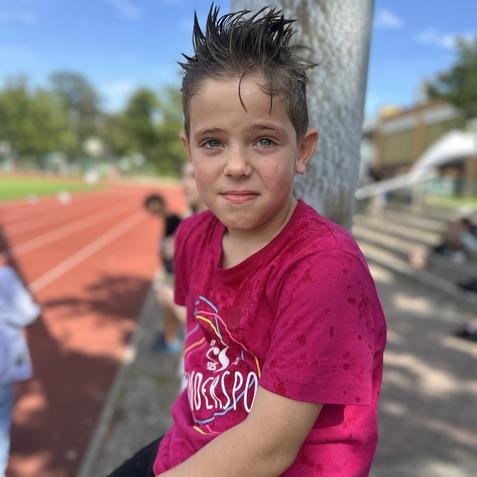 Junge mit nassem, hochstehendem Haar tr&auml;gt ein rotes T-Shirt, sitzt an einem Sportplatz mit Laufbahn im Hintergrund.
