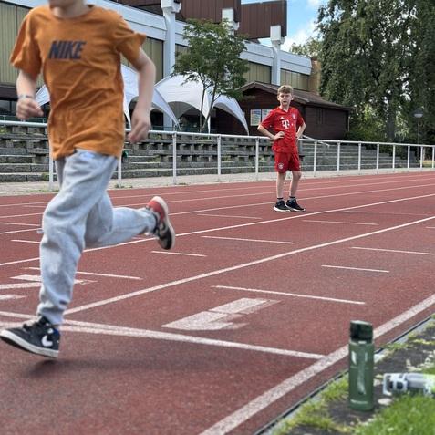 Zwei Jungen laufen auf einer roten Laufbahn; einer sprintet, w&auml;hrend der andere steht und zusieht.