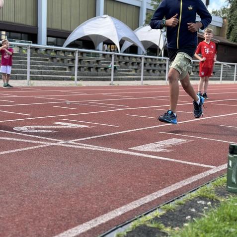 Ein Junge l&auml;uft auf einer roten Tartanbahn, im Hintergrund stehen Zelte und weitere Kinder.