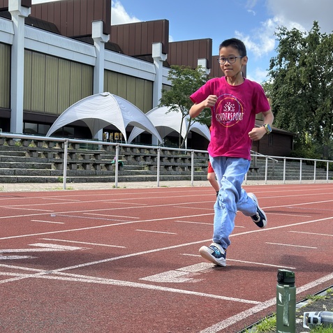 Ein Junge in pinkem Shirt l&auml;uft auf einer roten Laufbahn, im Hintergrund sind Zelte und ein Geb&auml;ude sichtbar.