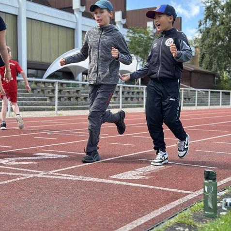 Zwei Jungen laufen fr&ouml;hlich auf einer roten Laufbahn, im Hintergrund sind weitere Kinder und eine Sporthalle sichtbar.