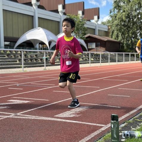 Ein Junge in pinkem Shirt l&auml;uft auf einer roten Laufbahn, im Hintergrund sind Sportgeb&auml;ude und B&auml;ume zu sehen.