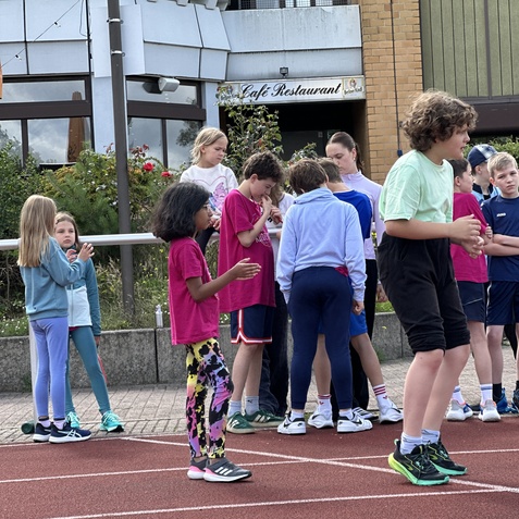 Gruppe von Kindern in sportlicher Kleidung versammelt auf einer roten Laufbahn, im Hintergrund ein Caf&eacute;.